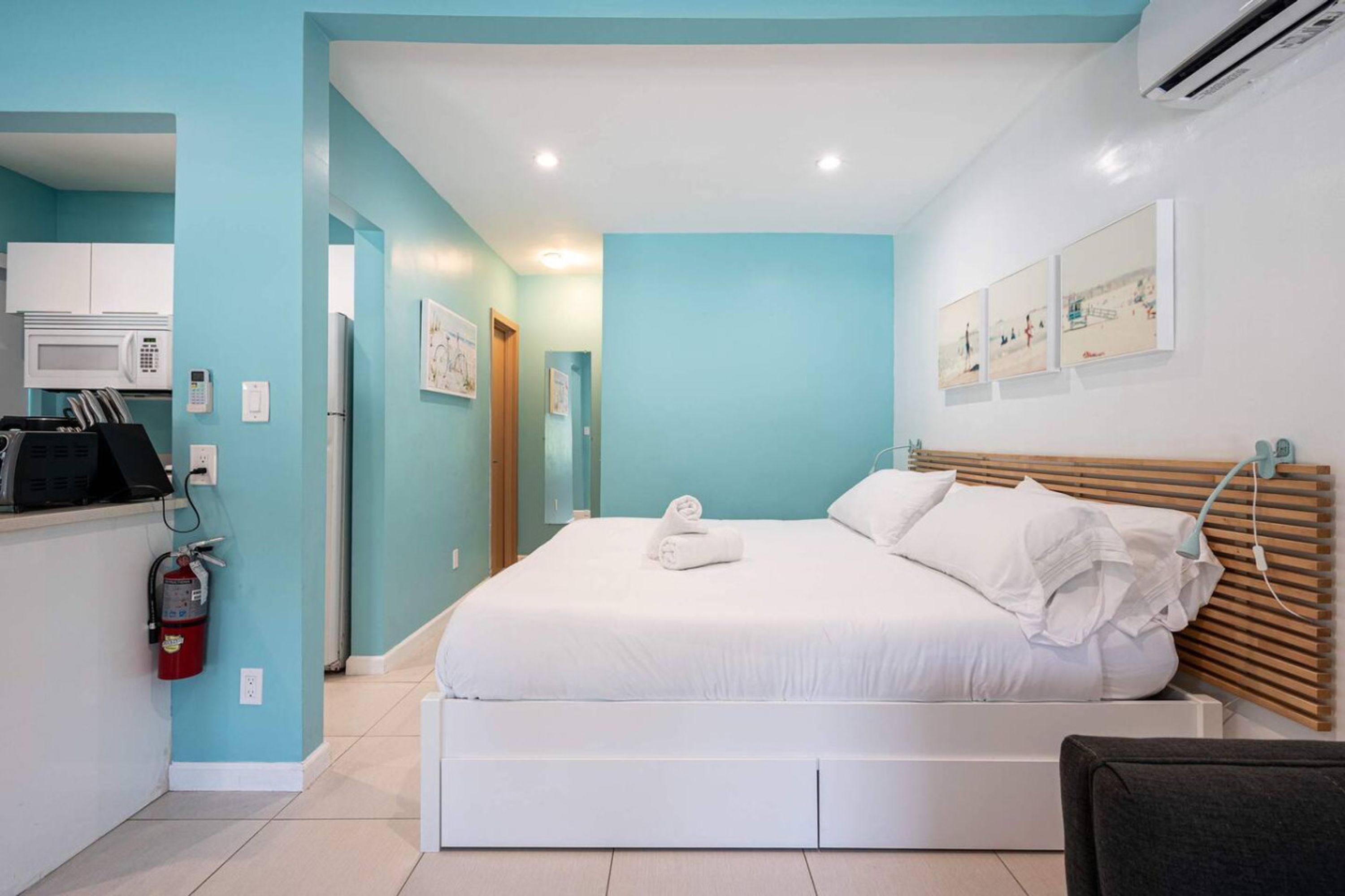 Quarto de hotel moderno com cama, escrivaninha com computador, micro-ondas e extintor de incêndio.