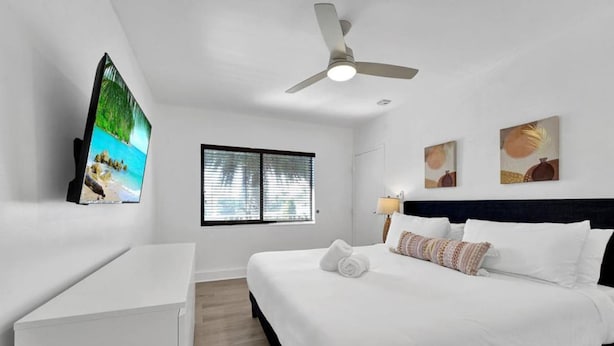 Una habitación de hotel con una cama, ventilador de techo, televisión y obras de arte en la pared.