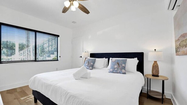 A modern bedroom with a large bed, a ceiling fan, a window with blinds, a bedside table with a lamp, and a painting on the wall. - Waves On Walnut 2-beach Access (Hollywood)