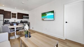 A modern kitchen with dark wood cabinets, a microwave, and a coffee maker. There is a dining table with chairs and a vase of flowers. A television is mounted on the wall, displaying a beach scene. - Waves On Walnut 2-beach Access (Hollywood)