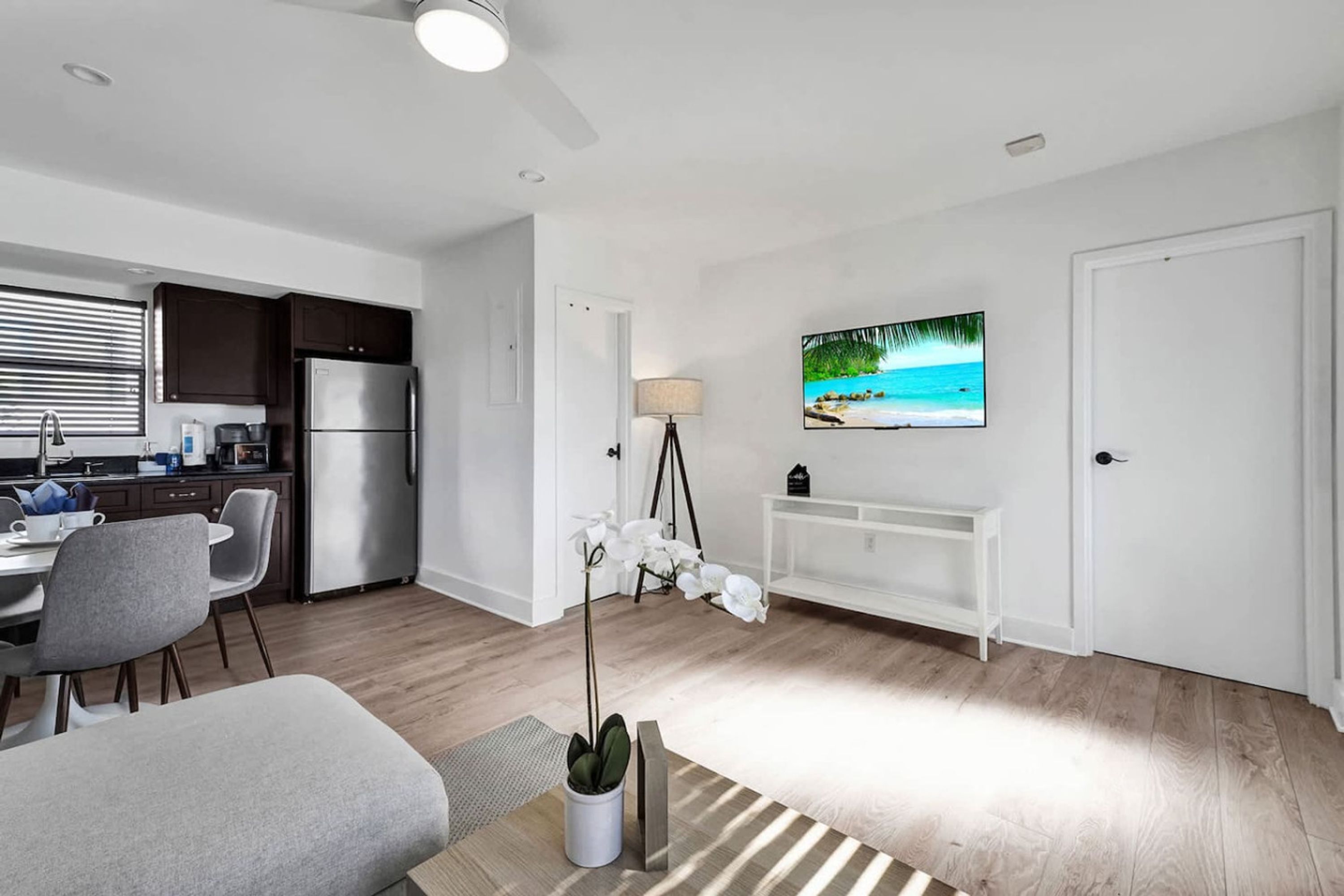 A modern kitchen with a refrigerator, stove, and dining area. There is a TV mounted on the wall and a potted plant in the living area.