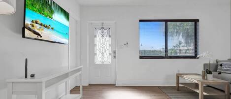 A modern living room with a large flat-screen TV on the wall, a wooden floor, a white door with decorative glass, and a window with blinds.