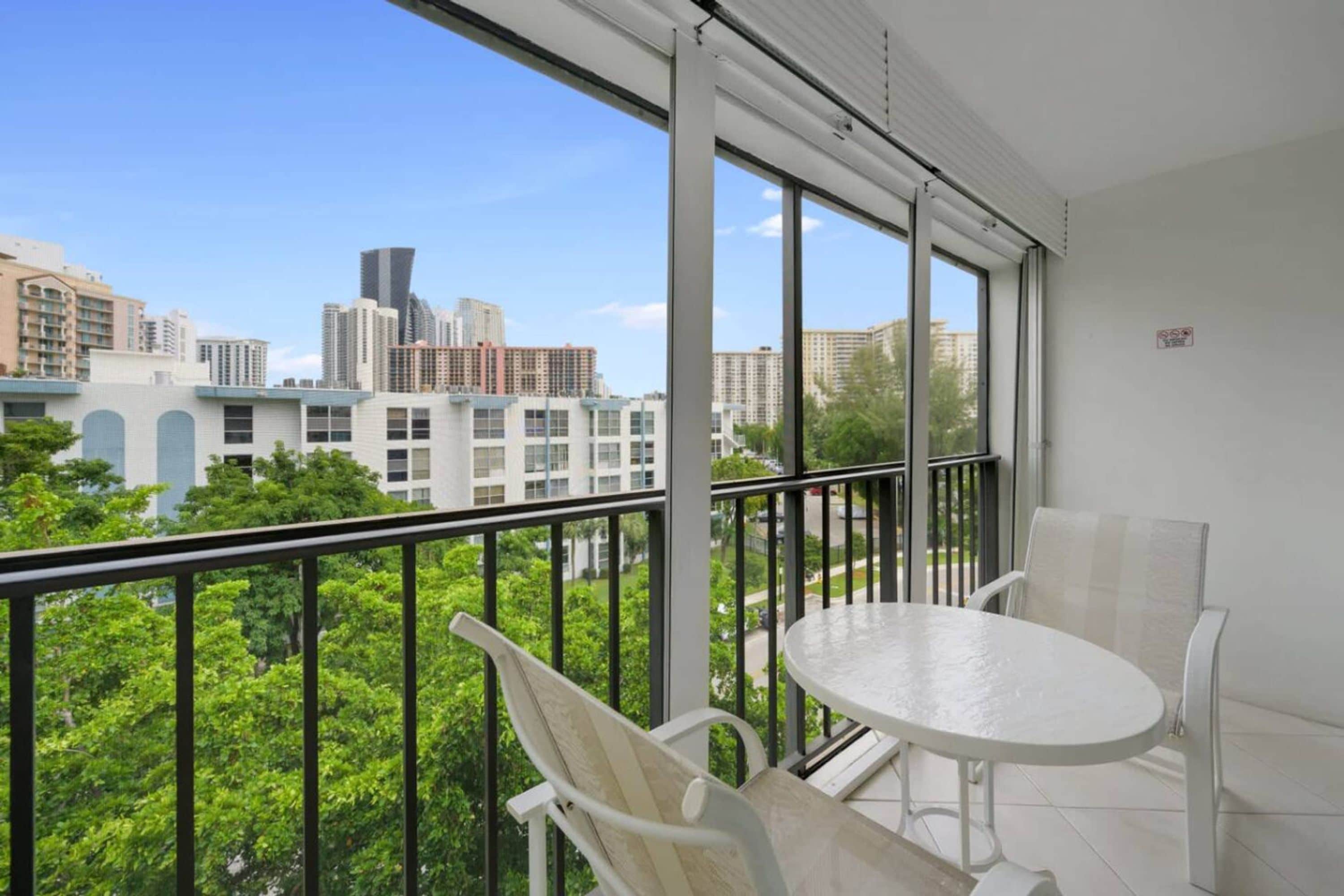A balcony with a white table and chairs, large windows offering a view of city buildings and greenery.