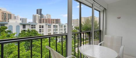 A balcony with a white table and chairs, large windows offering a view of city buildings and greenery.