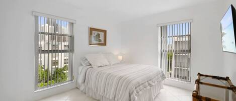 A bedroom with a bed, two windows with bars, a framed picture on the wall, and a wooden chair.
