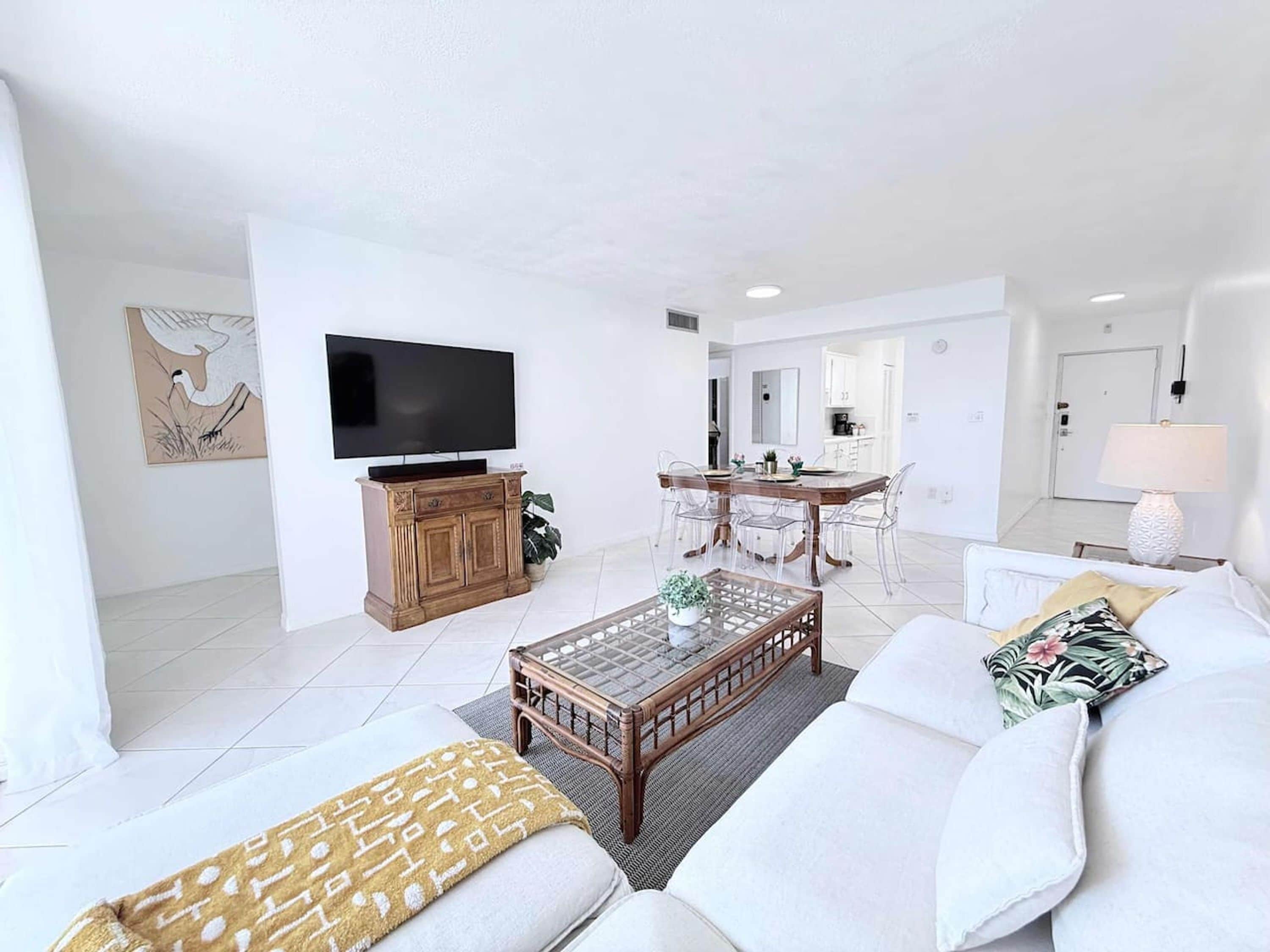 Een moderne woonkamer met een bank, salontafel en televisie. Er is een eethoek met stoelen en een tafel. De kamer heeft witte wanden en een betegelde vloer.