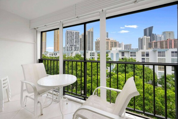 A balcony with white chairs and a table, offering a city view through large windows.
