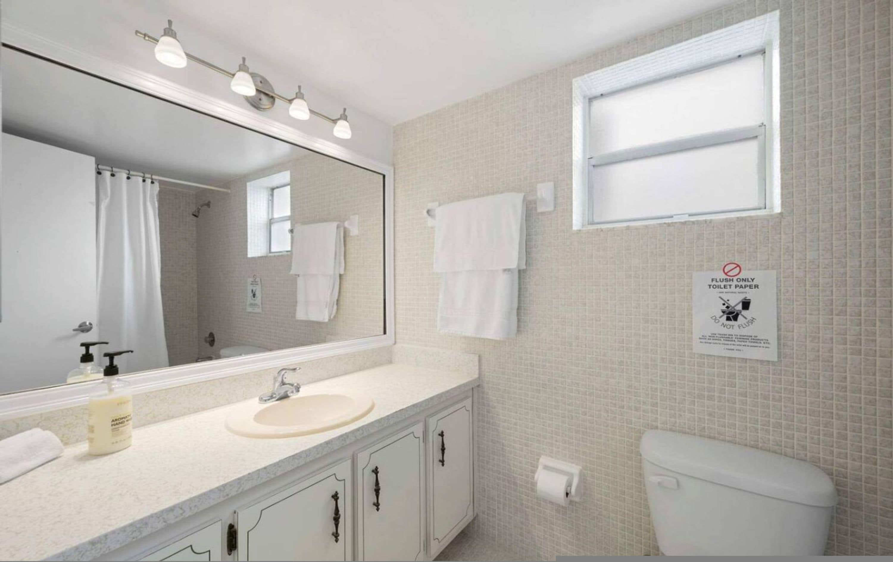 Une salle de bain avec un lavabo, un miroir et des toilettes. Un panneau sur le mur indique que les toilettes ne servent qu’à tirer la chasse d’eau.