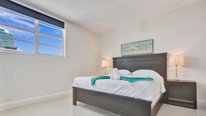 A bedroom with a large bed, two bedside tables with lamps, a window with a view of a building, and a painting on the wall. - New New Beach View (Hollywood)