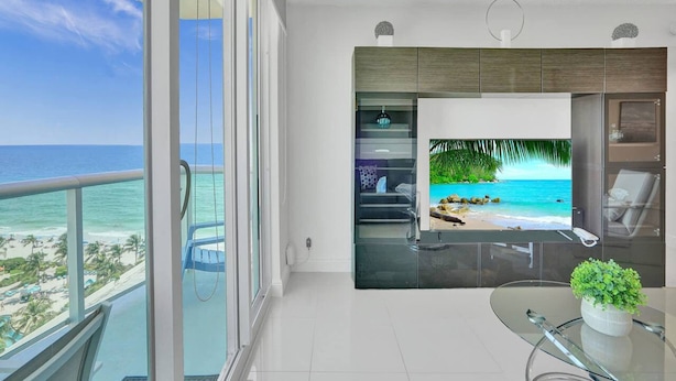 A modern living room with a large TV screen displaying a beach scene, a glass table with a plant, and a balcony with a view of the ocean. - New New Beach View (Hollywood)
