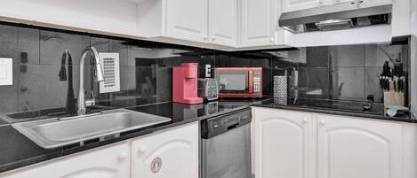Una cocina moderna con armarios blancos, encimera negra, pileta de acero inoxidable, cafetera roja, microondas y lavavajillas.