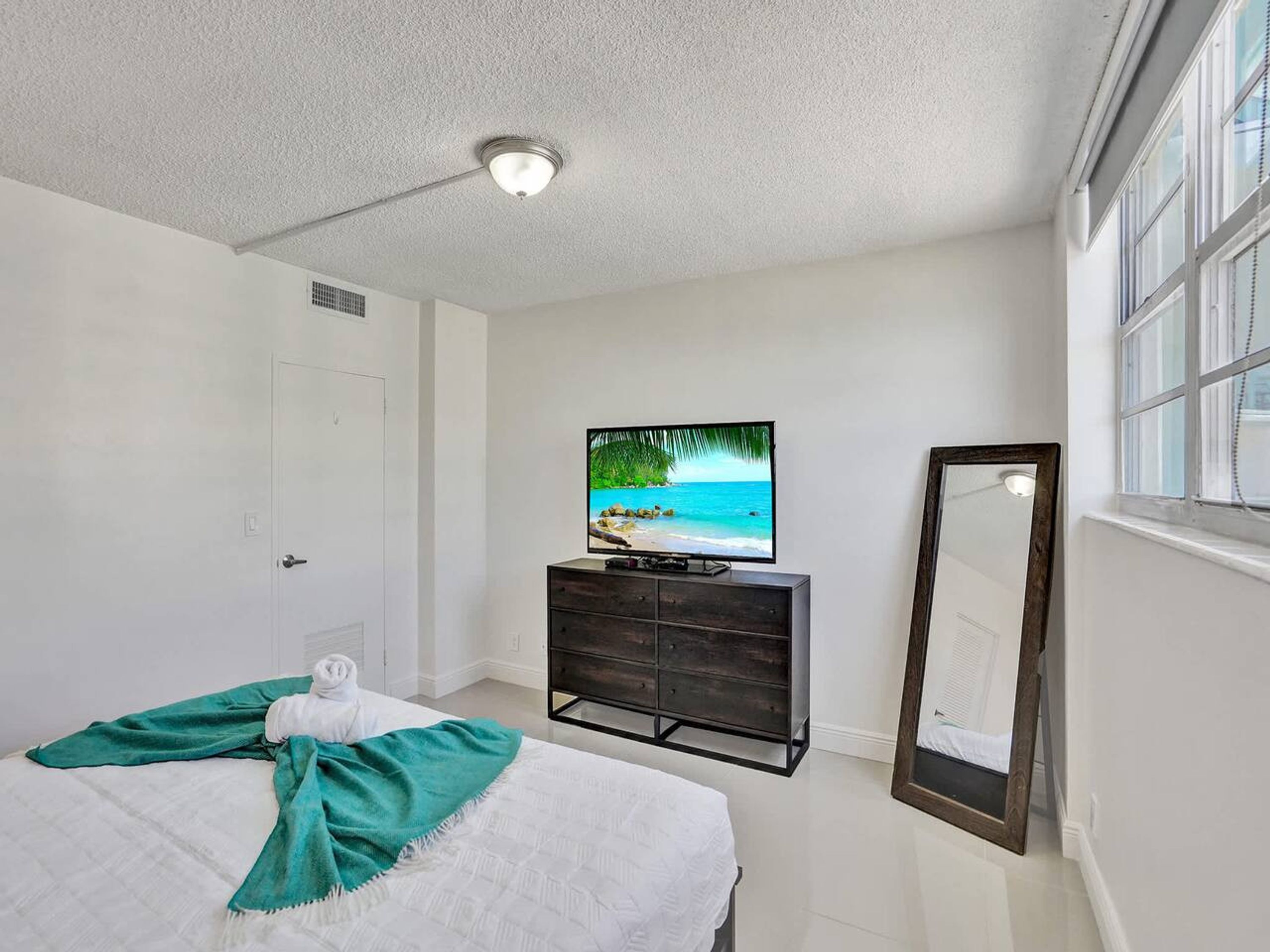 Een slaapkamer met een bed, een televisie op een dressoir, een raam en een spiegel.