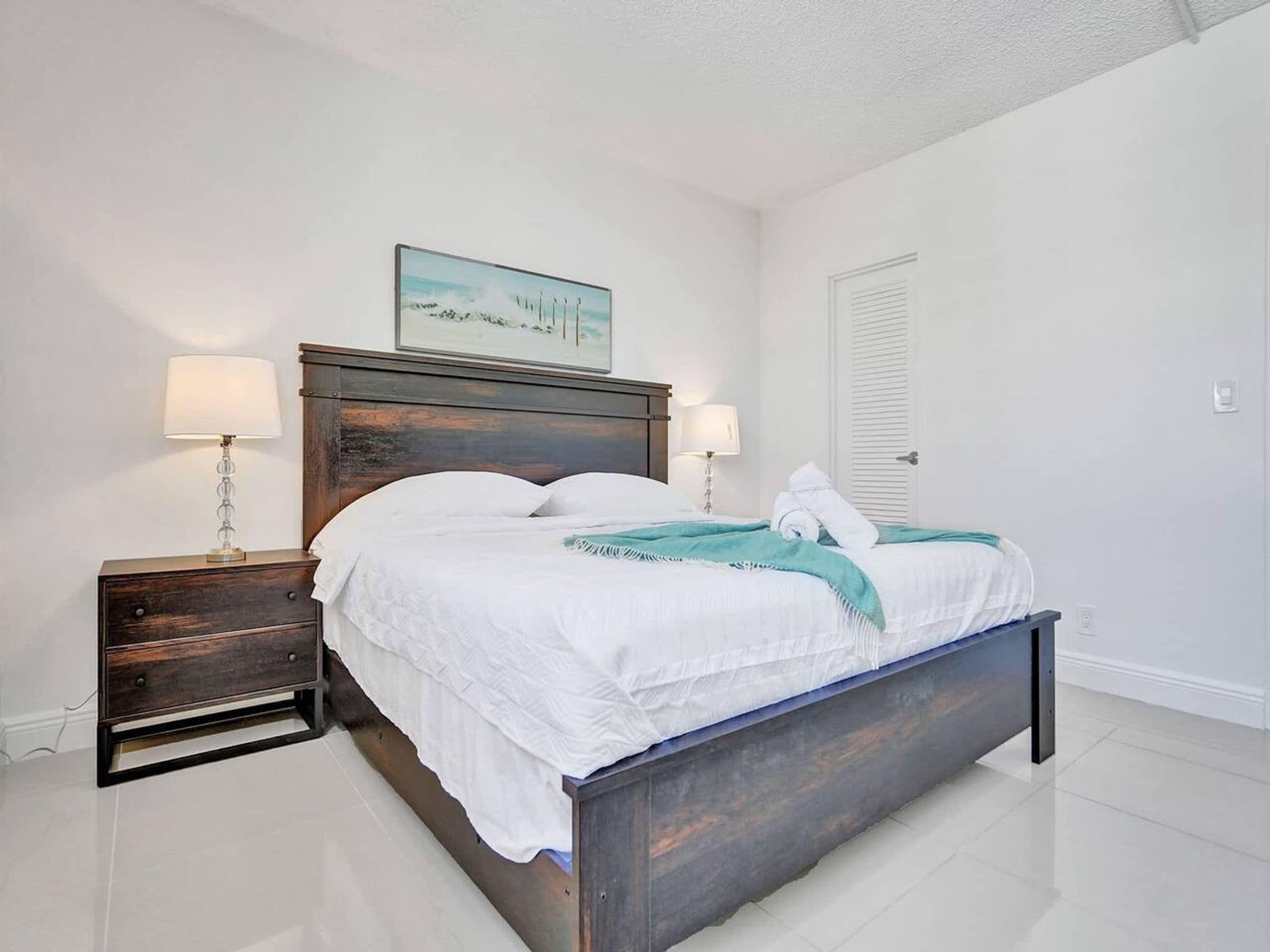 Un lit bien fait, avec du linge de lit blanc et un plaid turquoise, une table de chevet en bois avec une lampe, et un tableau encadré accroché au mur.