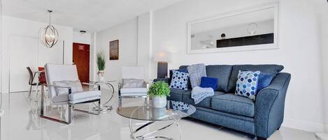Una sala de estar moderna con un sofá azul, una mesa de centro de vidrio transparente, paredes blancas y una lámpara de techo.