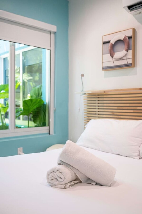A neatly made bed with a rolled white towel, a window with blinds, and a framed picture on the wall.