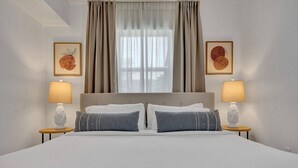 A hotel room with a large bed, two bedside tables with lamps, a window with curtains, and two framed pictures on the wall.