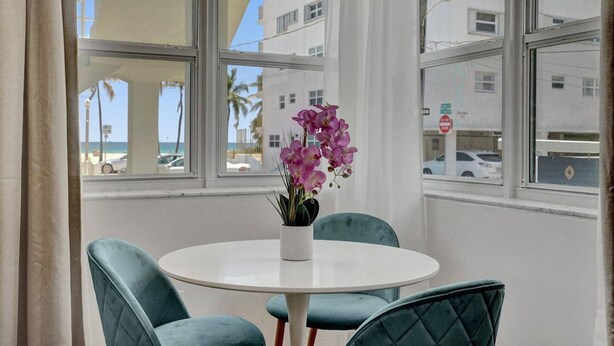 A room with a round table and two chairs, a potted orchid, and a view of buildings and palm trees through large windows.
