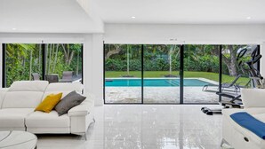 A spacious living room with a large window, a white sofa, and a view of a swimming pool and greenery outside. - Hibiscus House in Fort Lauderdale (Fort Lauderdale)