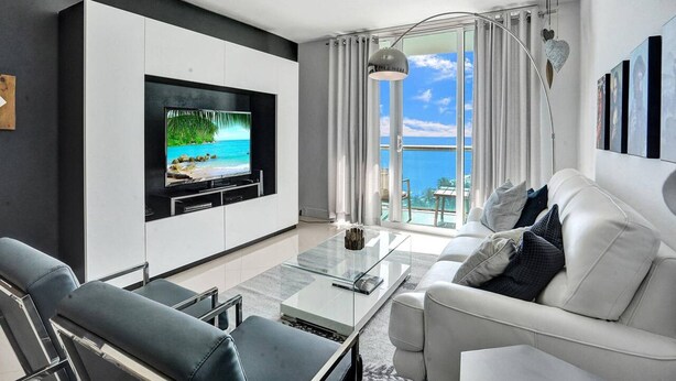 Una sala de estar moderna con un televisor de pantalla plana grande montado en un mueble blanco, una mesa de centro de vidrio, un sofá blanco con cojines y vistas al océano a través de amplias ventanas.