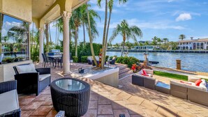 A covered outdoor patio with wicker furniture, a glass-top table, and a view of a water body with palm trees.