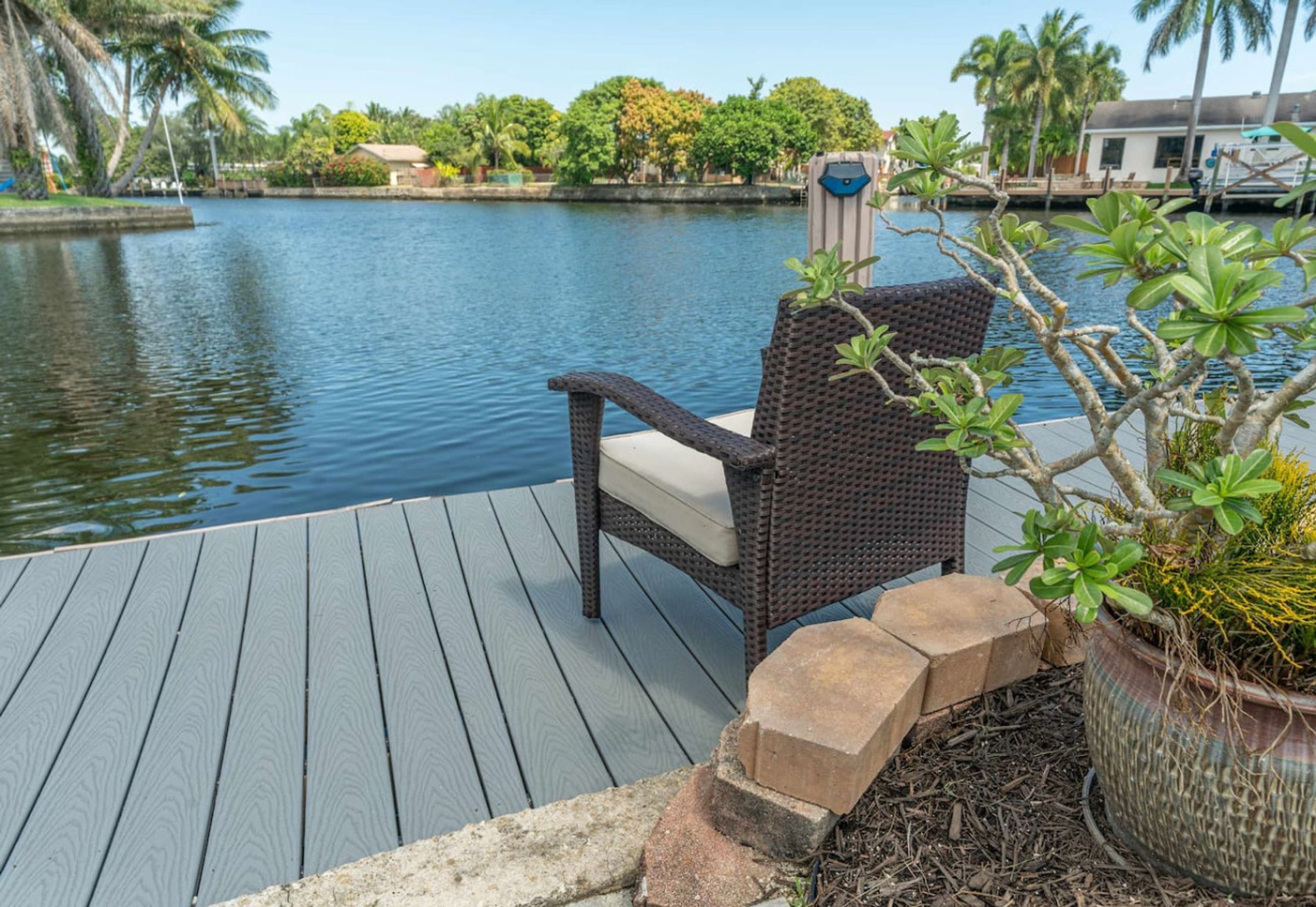 A wicker chair with a cushion on a wooden deck by a calm body of water, surrounded by lush greenery and a clear sky.