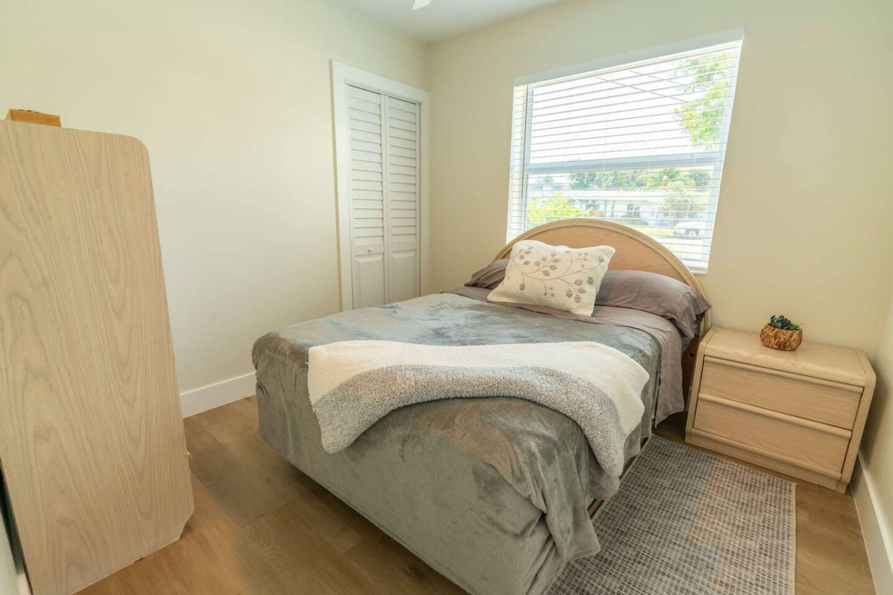A bedroom with a bed, a nightstand, a wooden wardrobe, a window with blinds, and a view of the outside.