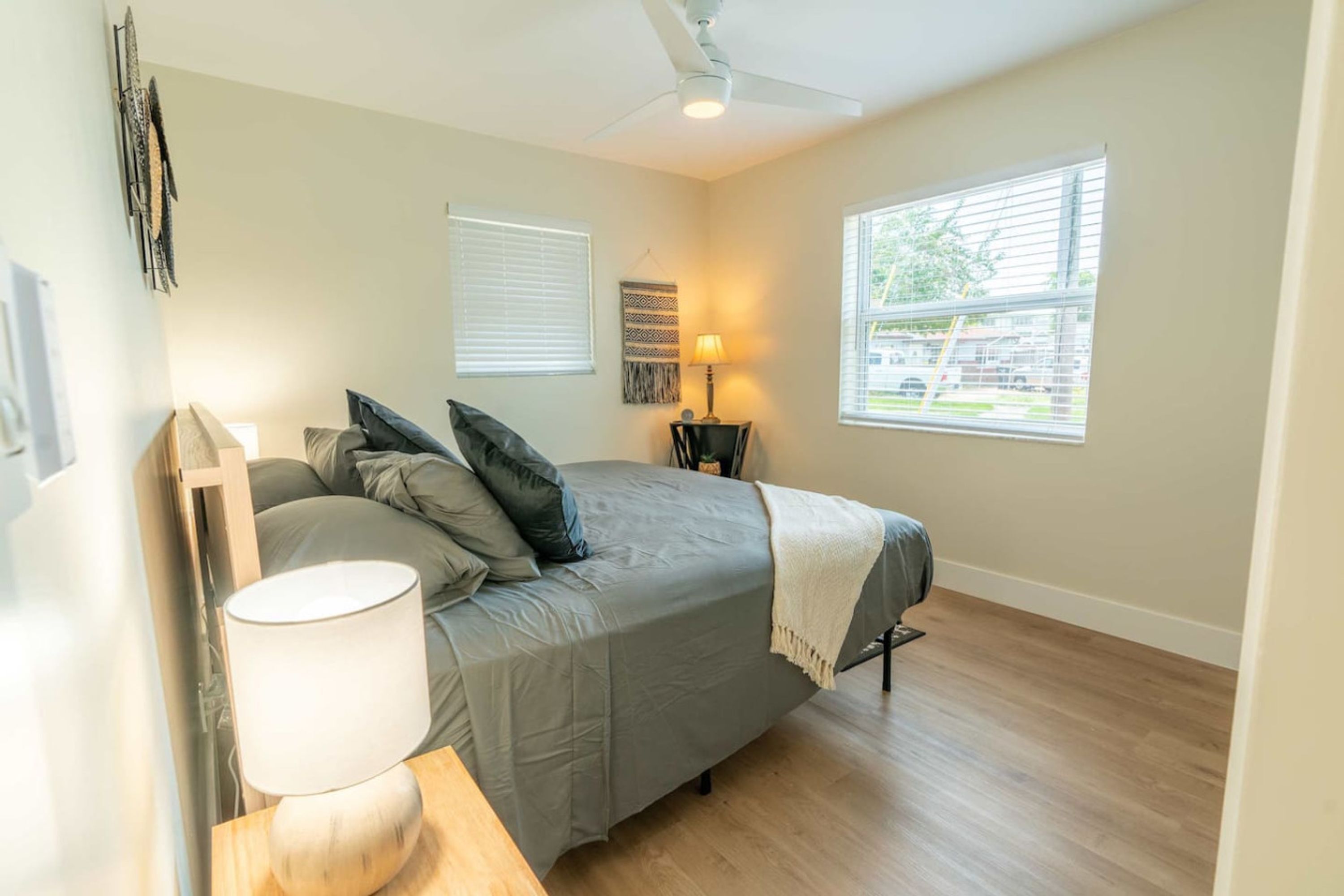 Une chambre à coucher avec un lit, une table de chevet avec une lampe, un ventilateur de plafond et une fenêtre avec des stores.
