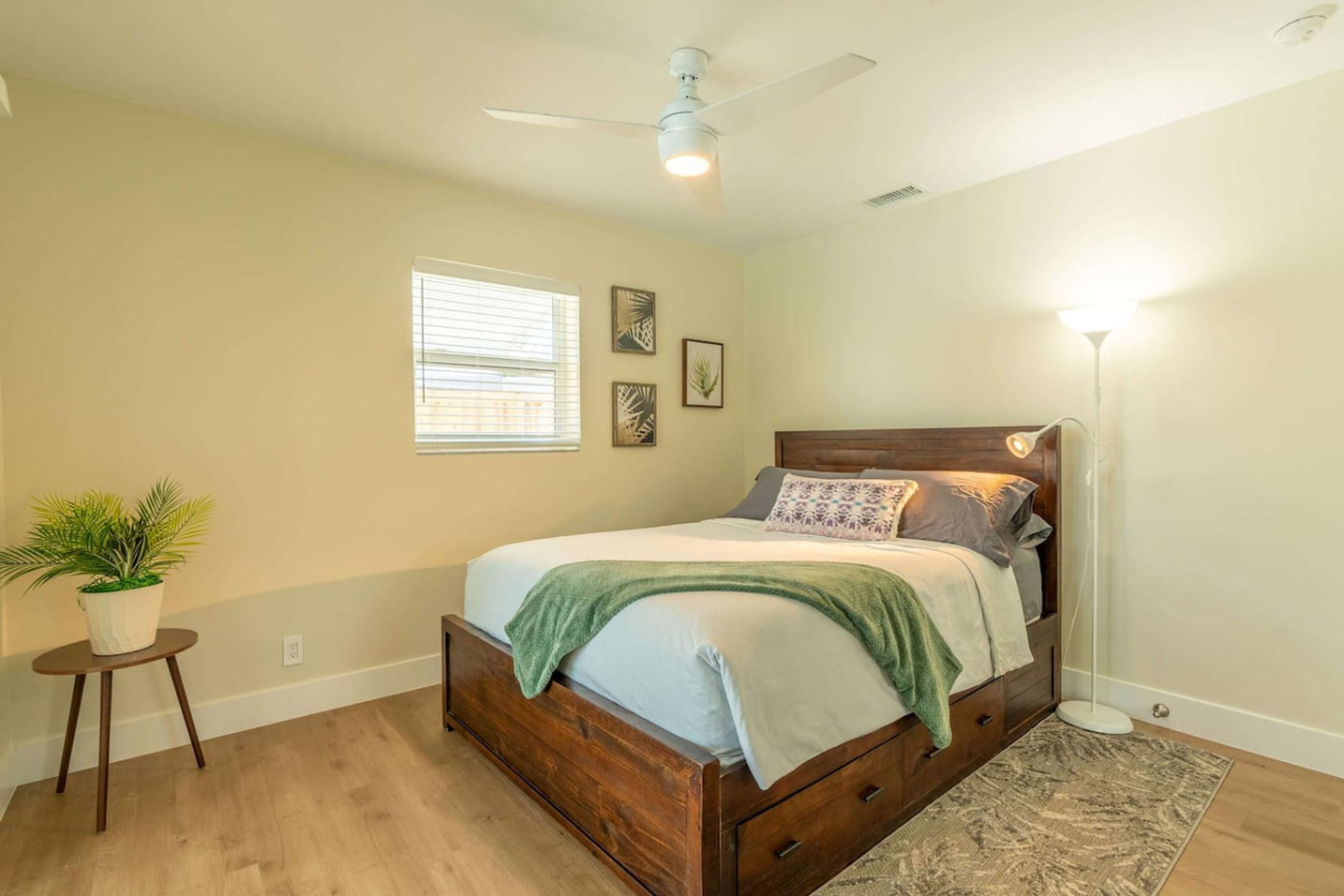 Un dormitorio con una cama de madera, un ventilador de techo, una lámpara de pie y una pequeña planta en maceta.