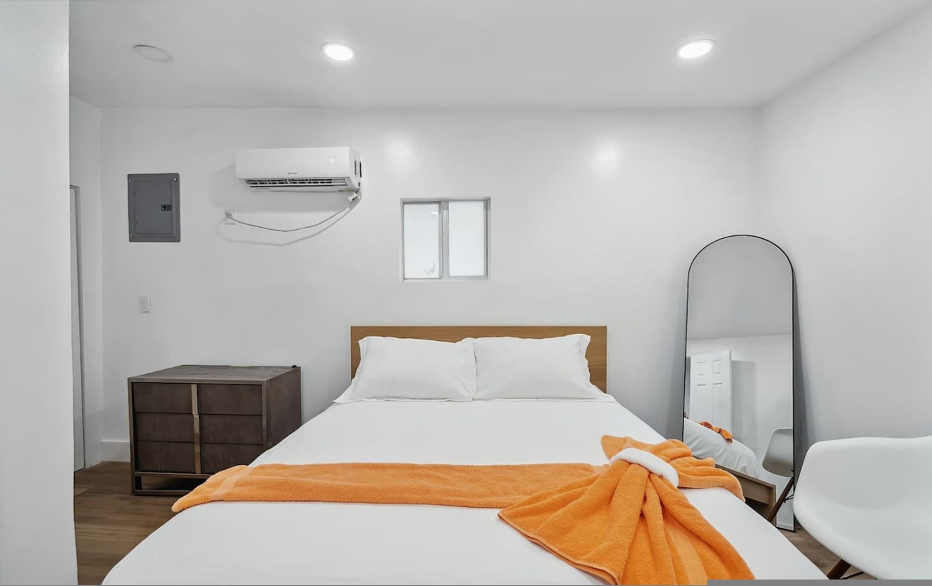 A hotel room with a bed, a bedside table, a wardrobe, an air conditioning unit, a window, and a mirror.