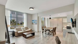 A modern living room with a sofa, coffee table, dining table, and chairs. There is a TV mounted on the wall, a window with curtains, and a view of buildings outside. - NEW NEW Ocean Oasis (Hollywood)