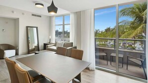 A modern hotel room with a large balcony, a dining area with a table and chairs, and a large window offering a view of palm trees and a parking lot. - NEW NEW Ocean Oasis (Hollywood)