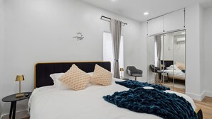 Ein modernes Schlafzimmer mit einem großen Bett, einem Nachttisch mit Lampe, einem Sessel, einem Spiegel und Blick ins Freie.