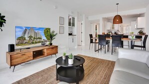 Una sala de estar moderna con un televisor de pantalla plana, un mueble de entretenimiento de madera, una mesa de centro negra y un comedor con una cocina al fondo.