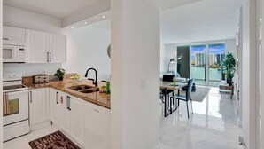 Una cocina moderna con armarios blancos, encimera de granito y microondas integrado. Cuenta con un comedor con mesa y sillas, y vistas a un paisaje urbano a través de amplios ventanales.