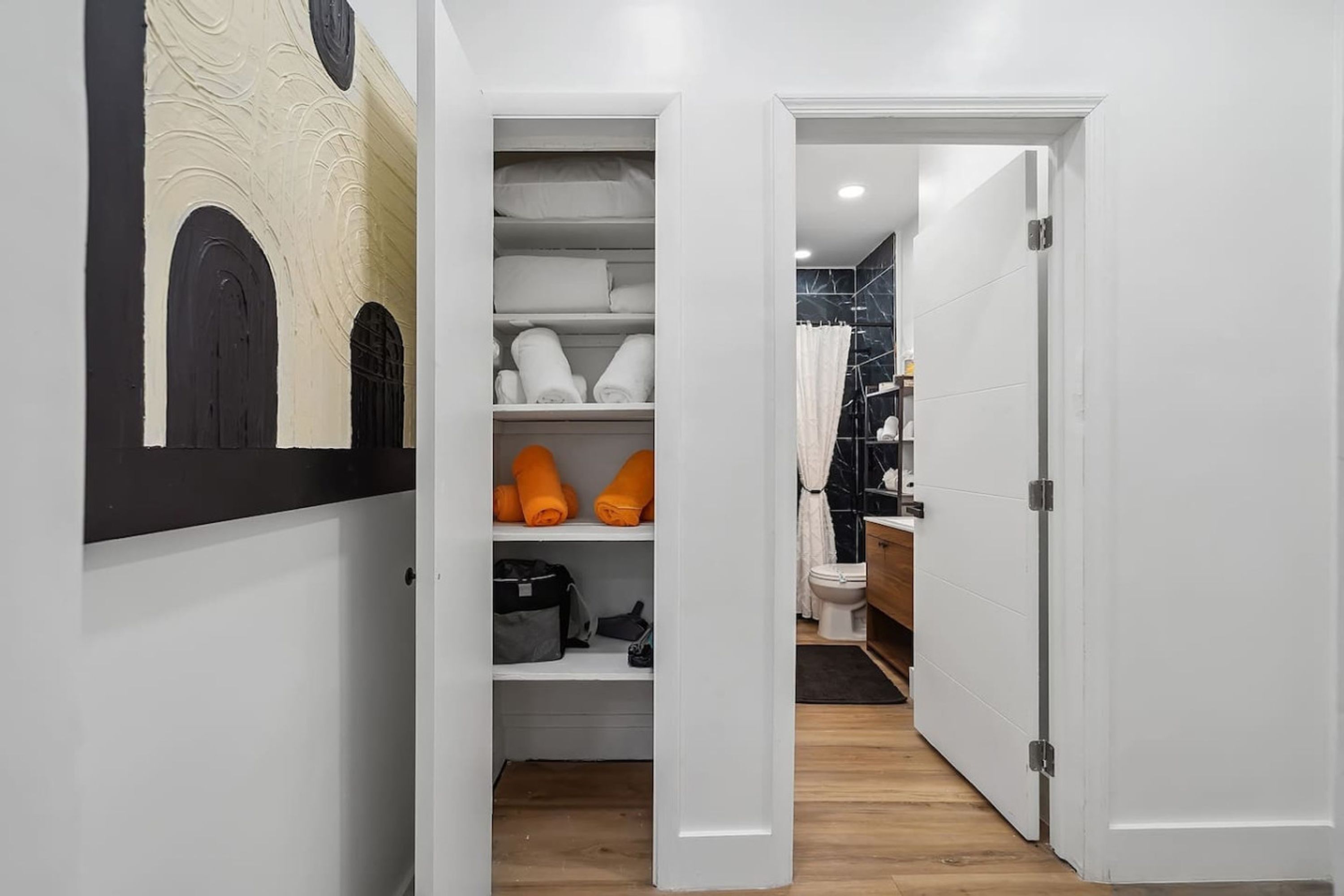 Un pasillo estrecho con una puerta blanca que da a un baño. La pared, de color blanco, presenta una obra de arte en blanco y negro. Hay estantes con toallas dobladas y un bolso negro.