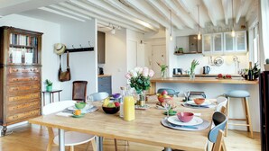 Uma sala de jantar com uma mesa de madeira posta para seis lugares, cercada por cadeiras. Um armário de madeira com gavetas fica à esquerda.