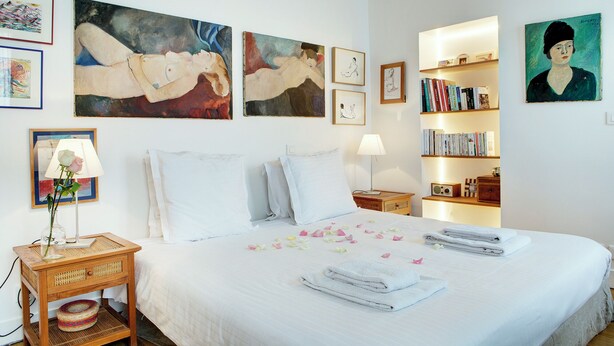Quarto de hotel com uma cama grande, uma mesa de cabeceira com abajur, uma mesinha lateral com um vaso de flores e uma estante de livros.