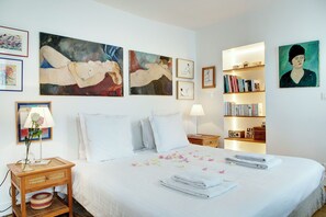 Una habitación de hotel con una cama grande, una mesita de noche con lámpara, una pequeña mesa auxiliar con un jarrón de flores y una estantería.