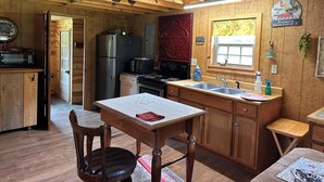 A rustic cabin interior with wooden walls, a kitchen area including a refrigerator, stove, and microwave, a dining table with chairs, and a window with a view.