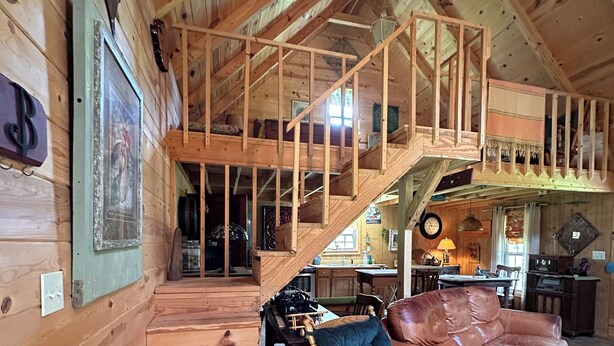 A wooden cabin interior with a loft bedroom, a dining area, and a living room with a brown leather sofa and a blue armchair.