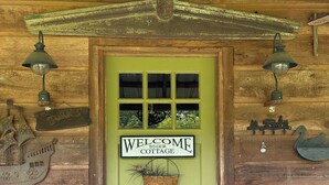 A wooden cottage with a green door, a welcome sign, and rustic decor including a bench and a birdcage.