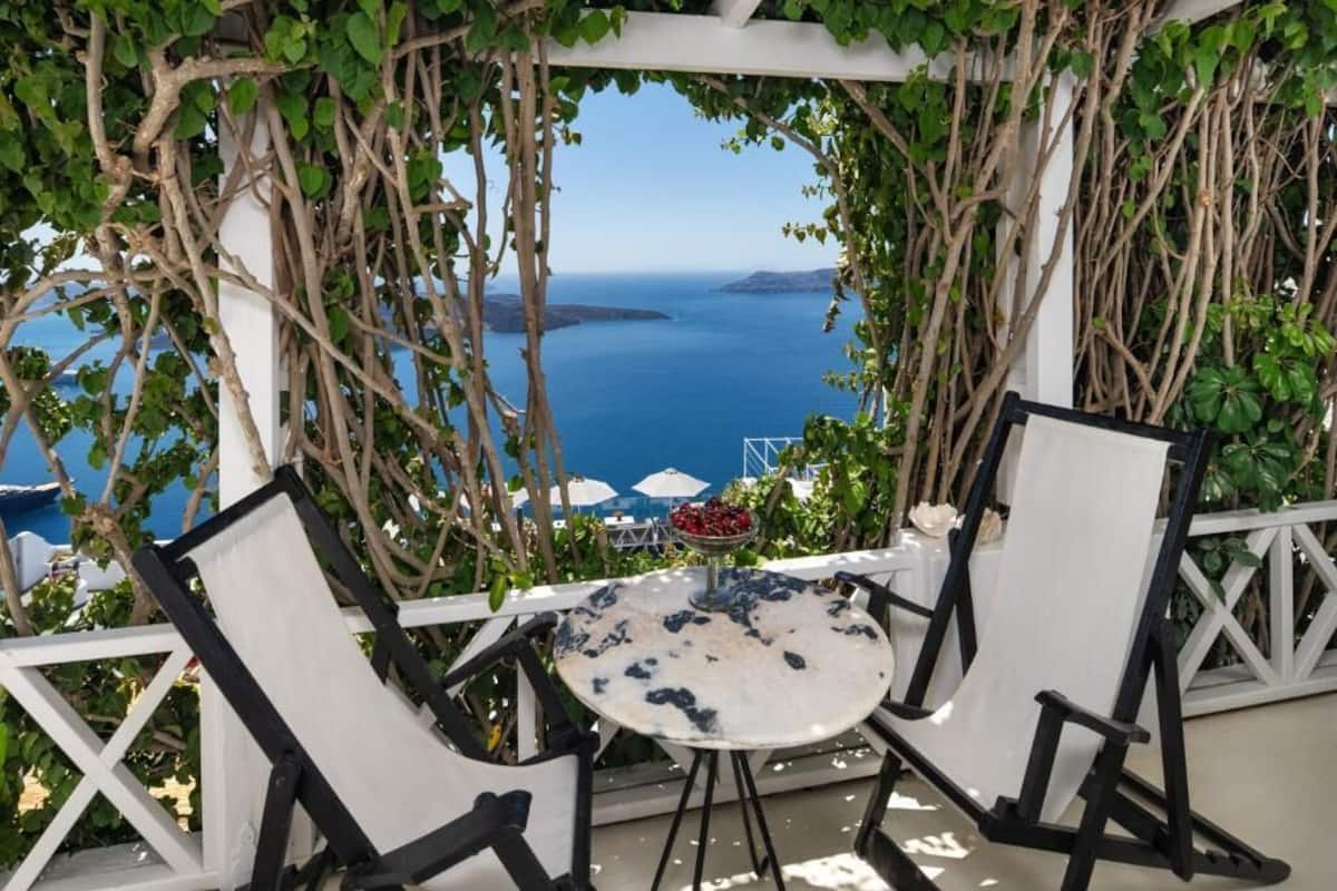 Une table avec deux chaises, placée sous une pergola couverte de vignes, avec vue sur un plan d’eau.