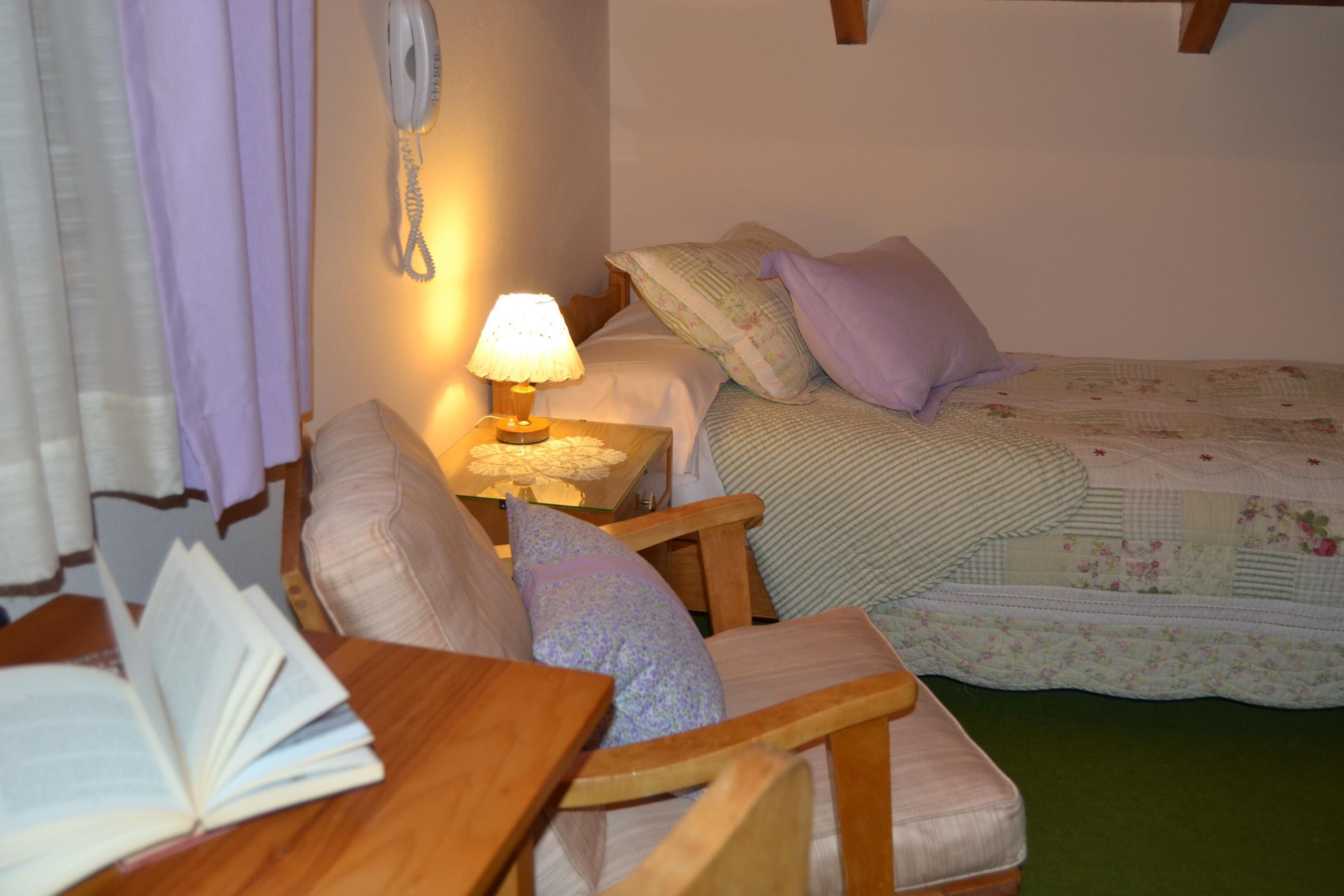 A cozy bedroom with a wooden chair, a bedside table with a lamp, a bed with floral bedding, and a telephone on the wall.