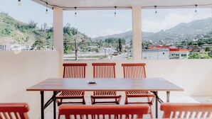 Uma varanda com mesa e cadeiras, com vista para uma paisagem montanhosa com edifícios e uma construção de telhado vermelho.