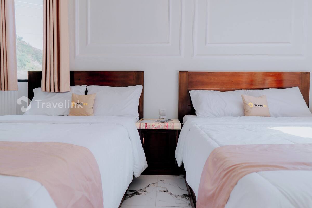 Een hotelkamer met twee bedden, elk met een houten hoofdbord, wit bedlinnen en een klein nachtkastje met een lamp.