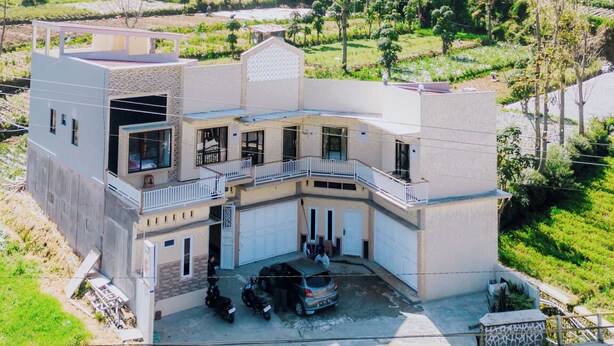 Um edifício de dois andares com telhado plano, varanda e garagem. Localiza-se numa área montanhosa com campos em socalcos ao fundo.