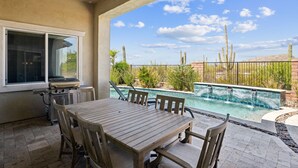 Um pátio externo com mesa e cadeiras de madeira, churrasqueira e piscina com cascata.