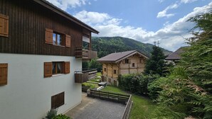 A traditional chalet-style building with a balcony, surrounded by lush greenery and mountains in the background.