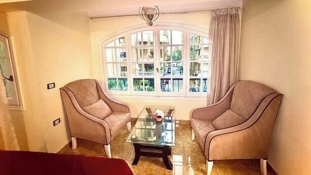 Une pièce avec deux fauteuils placés face à une grande fenêtre en arc de cercle, une table basse en verre et un luminaire suspendu au plafond.