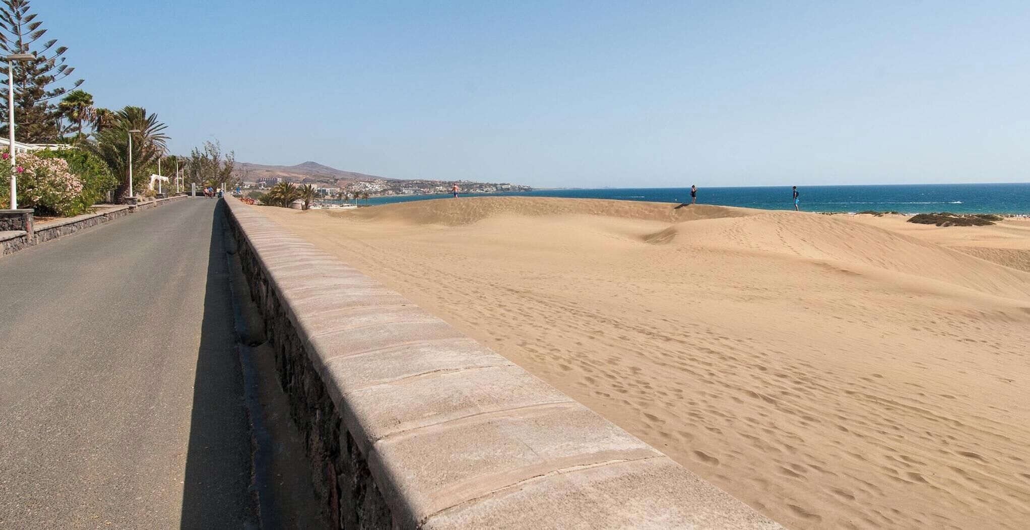A coastal road with a concrete barrier, sandy dunes, and a clear view of the sea.